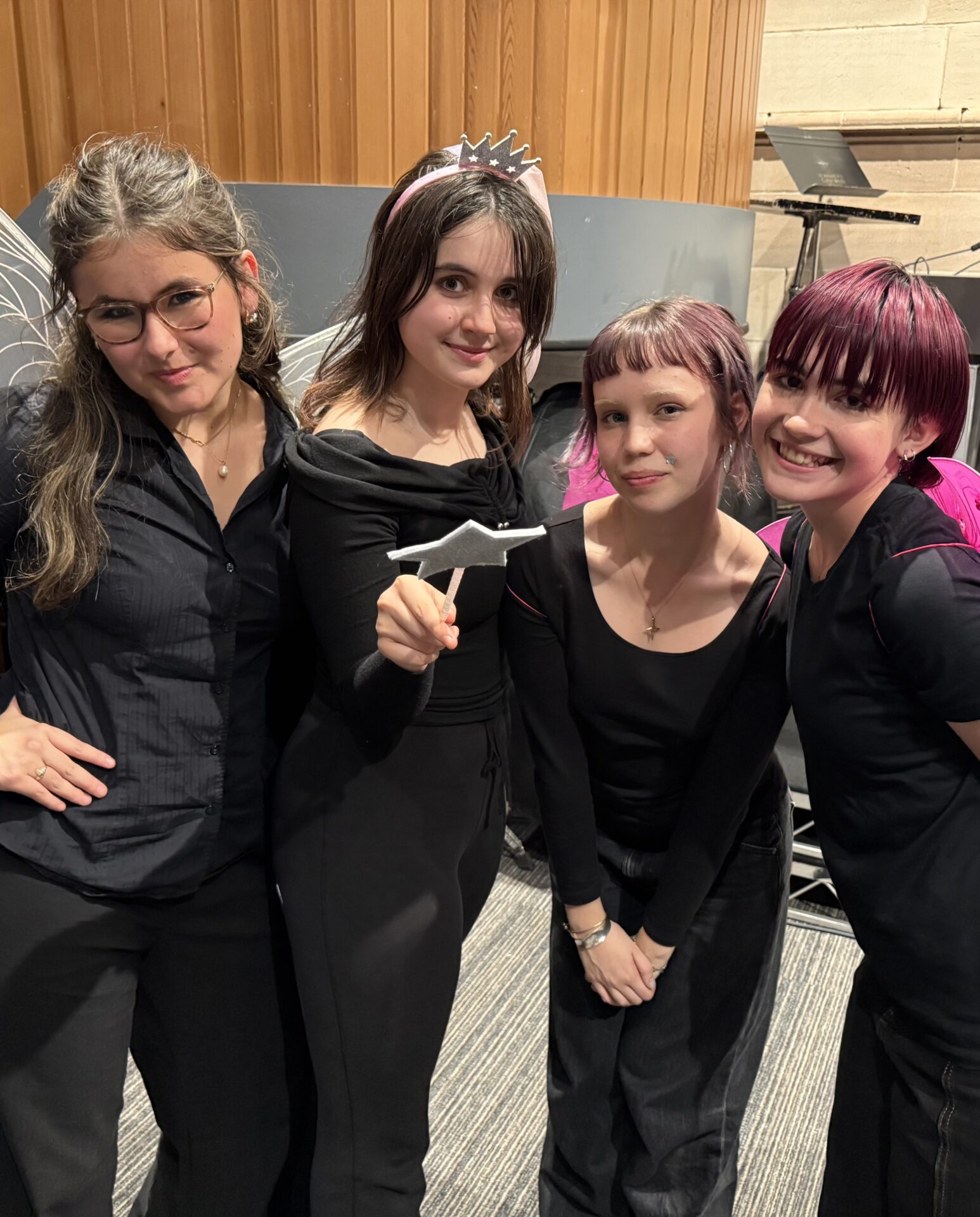 Four young women stand indoors, smiling at the camera in their black outfits. One, wearing a crown headband and holding a white origami bird, celebrates reaching the Theatresports Semi-Finals with her IGS teammates.