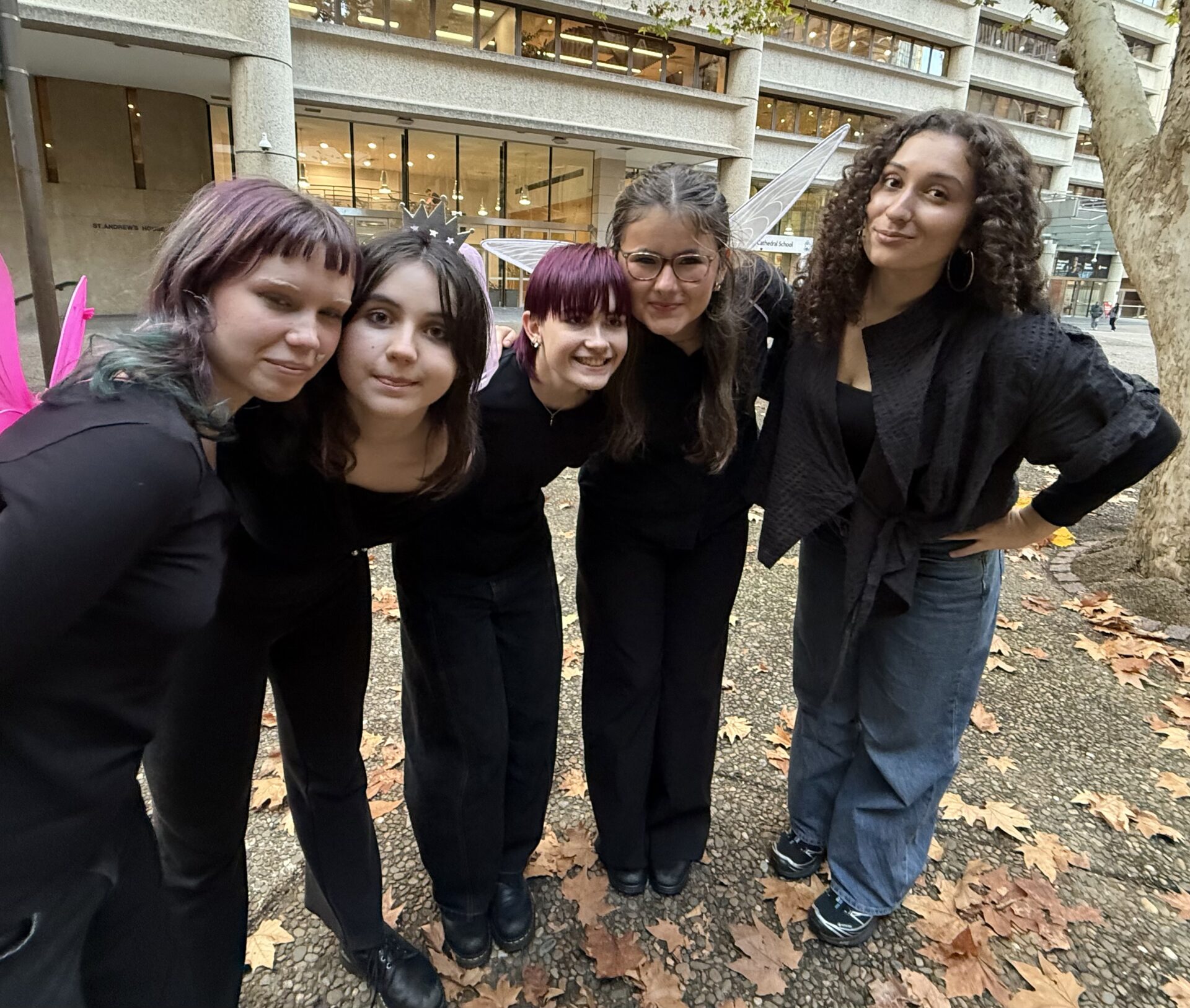Five young women stand close together outdoors, smiling at the camera. Dressed in dark clothing and surrounded by autumn leaves, they celebrate their IGS Theatresports Semi-Finals achievement with a modern building in the background.