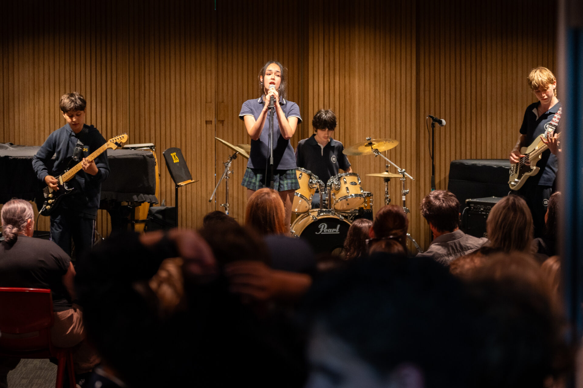 A school band performs on stage in front of an audience. Four pupils play guitar, bass, and drums while one sings into a microphone, with wooden panels as the backdrop.
