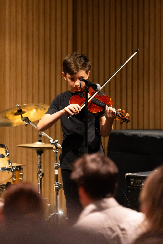 A young boy in a black outfit plays the violin on stage in front of an audience, with a drum kit and an amplifier visible in the background.