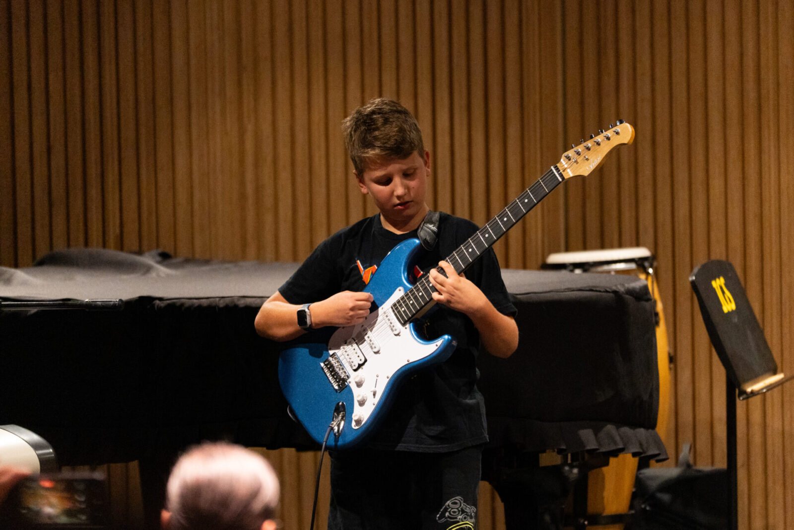 A young boy stands on stage playing a blue electric guitar, focused on his performance. A piano and percussion instruments are visible in the background; the setting appears to be a music recital or concert.