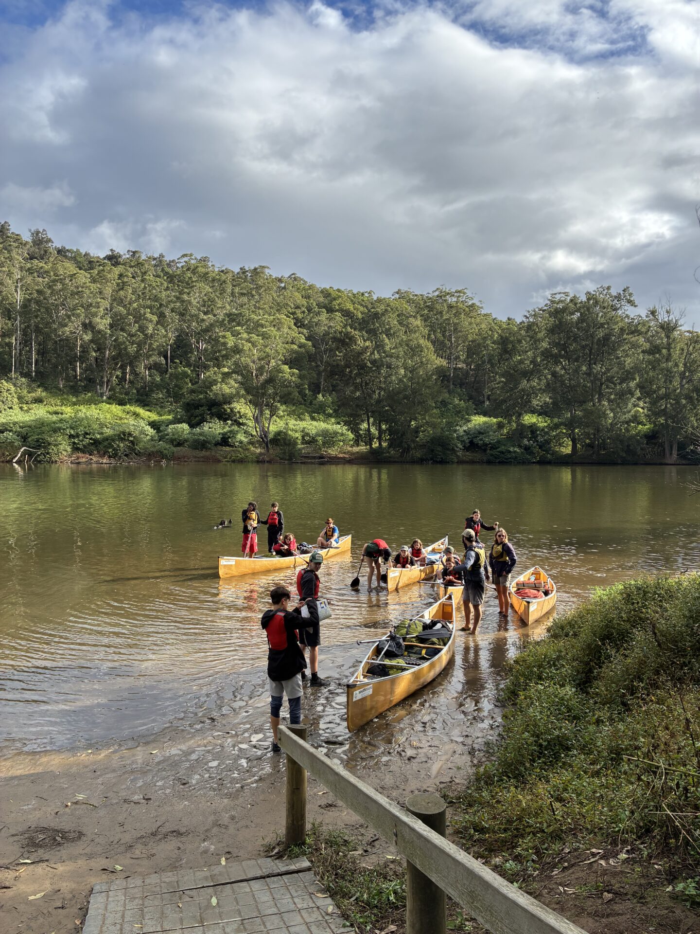 A group of people wearing life jackets stand near three yellow canoes at the edge of a calm river, surrounded by green trees and a cloudy sky, ready for an adventure into the valley. Some are in the shallow water, preparing or finishing a canoe trip.