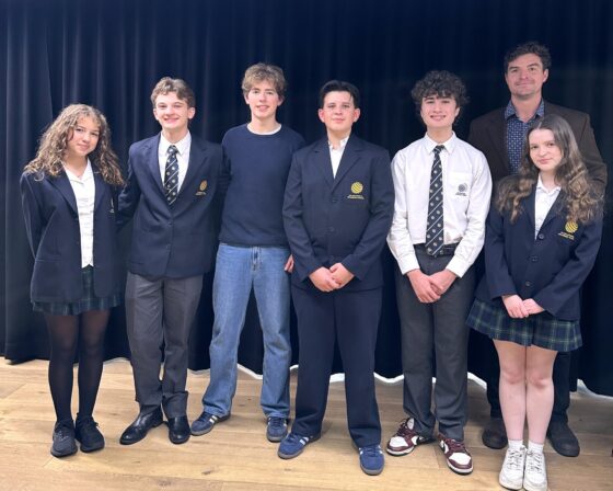 Seven people stand in a row on a wooden floor in front of a black curtain. Six teenagers in school uniforms or casual clothes are joined by an adult in a brown jacket, all smiling and facing the camera, celebrating their recent legal victory.