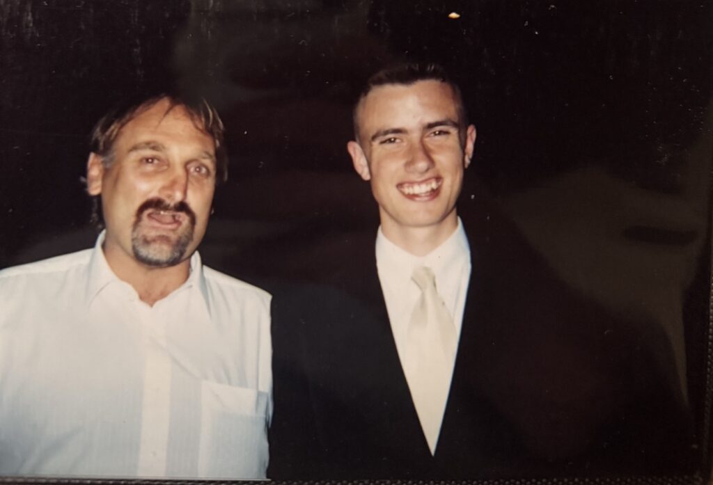 Two men are standing together and smiling at the camera. The man on the left, WITWAT’s Gabriel Shipton, has a beard and wears a light-coloured shirt, while the younger man on the right sports a dark suit and light tie. The background is dark.