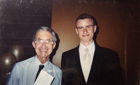Two men stand side by side, smiling at the camera. Gabriel Shipton wears a dark suit and light tie, while the older man in glasses holds a paper. WITWAT balloons and a brown wall decorate the background.