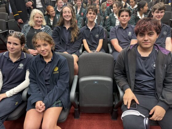 A group of students in navy blue and dark green uniforms, future leaders promoting gender equality, sit and smile in rows of grey theatre seats alongside a few adults in an auditorium setting.