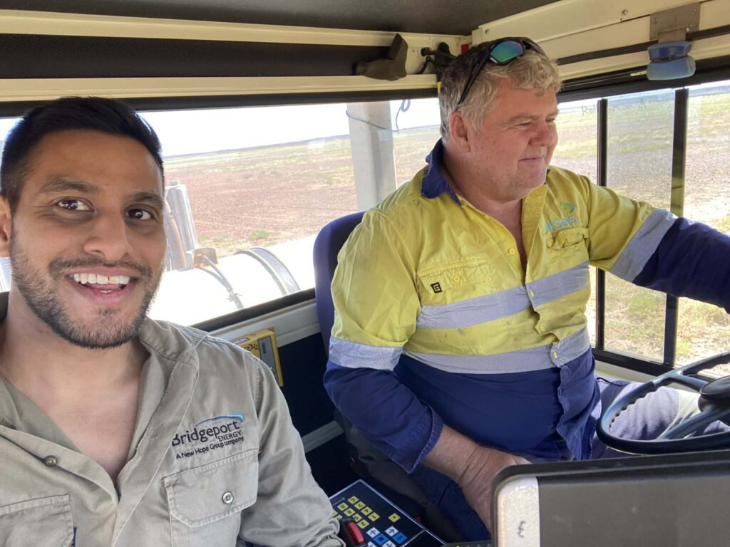 Two men wearing work shirts sit inside a vehicle. One smiles at the camera, while the other drives. Bright daylight streams through the windows, illuminating their faces and revealing fields outside—perhaps on their way to a WITWAT project or documentary shoot with Gabriel Shipton.