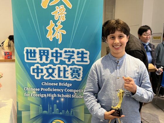 A smiling student in a light blue shirt holds a trophy, celebrating success whilst standing next to a banner for the Chinese Bridge Competition for Foreign Secondary School Students.