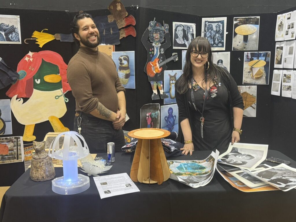 Two people stand smiling behind a table displaying art projects, including a glowing model and wooden structure, celebrating passion discovery amid colourful artwork, sketches, and photos covering the background wall.