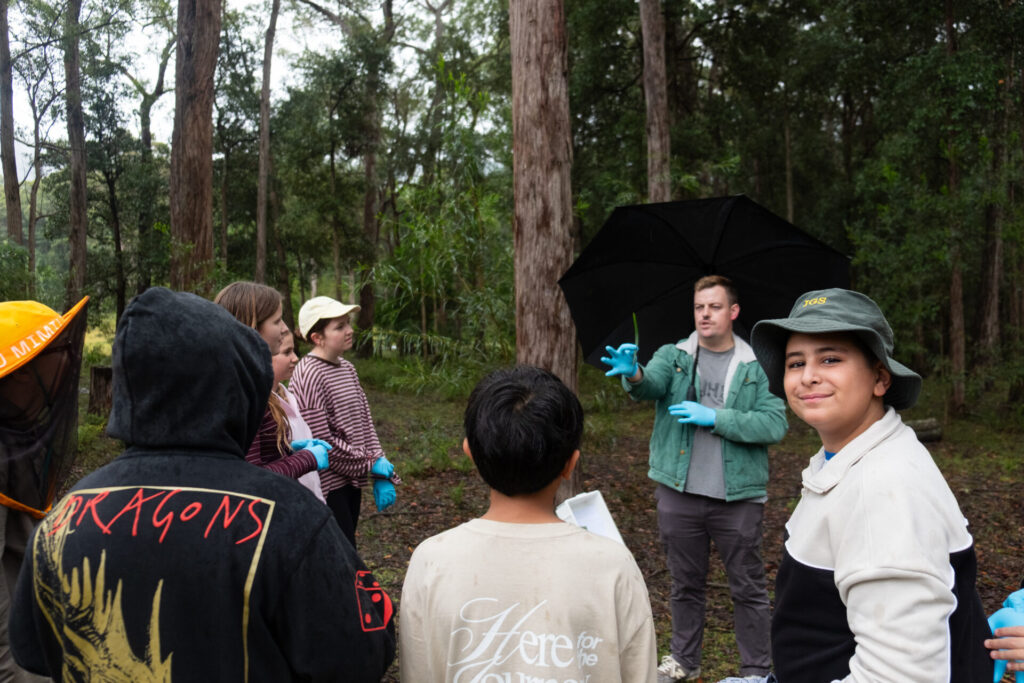 A group of children and a man wearing blue gloves gather in a forest for a Habitat Walk. The man holds an umbrella and gestures while talking. One boy in the foreground smiles at the camera as tall trees surround the group.