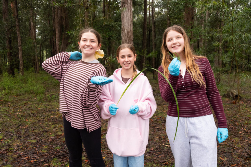 Three smiling girls stand in a forest during a Habitat Walk at the Retreat, wearing blue gloves. Two hold long green plants, and all are dressed casually in striped or light-coloured tops. The background is dense with trees and greenery.