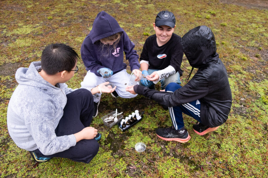 Four kids sit in a circle on grassy ground at the Retreat, wearing hoodies and jackets, as they examine scientific equipment and samples collected during their Habitat Walk. Small bottles and tools rest in a tray at the group's centre.