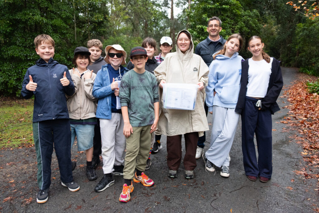 A group of smiling children and two adults stand on a wet path in a lush, green forest during a Habitat Walk. One adult holds a plastic box with papers inside. The group, dressed casually in hoodies and rain gear, enjoys this outdoor retreat.