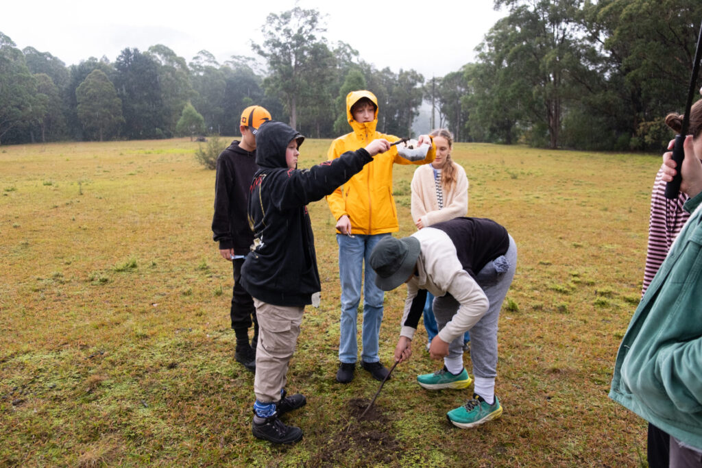 A group of young people stand in a grassy field, some wearing raincoats and hoodies. During their Habitat Walk Retreat, one points ahead whilst another digs in the ground. Trees and fog create a peaceful nature backdrop.