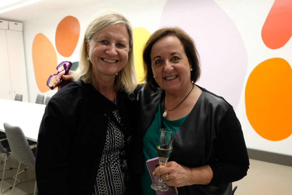 Two women smiling and posing together indoors, one holding a drink. Against a white wall with large, colourful abstract shapes, they appear happy and casually dressed—perfectly embodying the IGS spirit of designing a world-ready future.