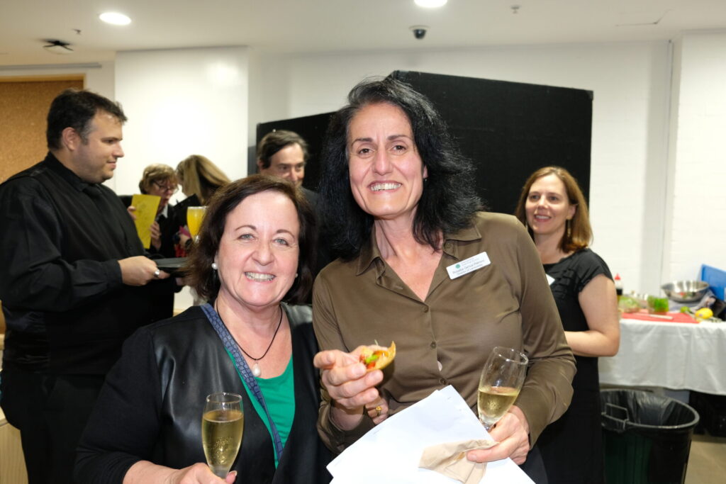 Two women smile at a social event, holding champagne and food. As others mingle by a table of drinks and nibbles, the friendly atmosphere reflects an IGS community designing world-ready experiences together.
