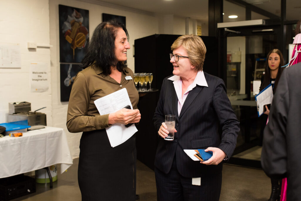 Two women stand indoors having a conversation; one holds papers and wears an IGS name badge, the other is in a suit holding a drink. There are tables, a glass door, and some people in the background at this World-Ready event.