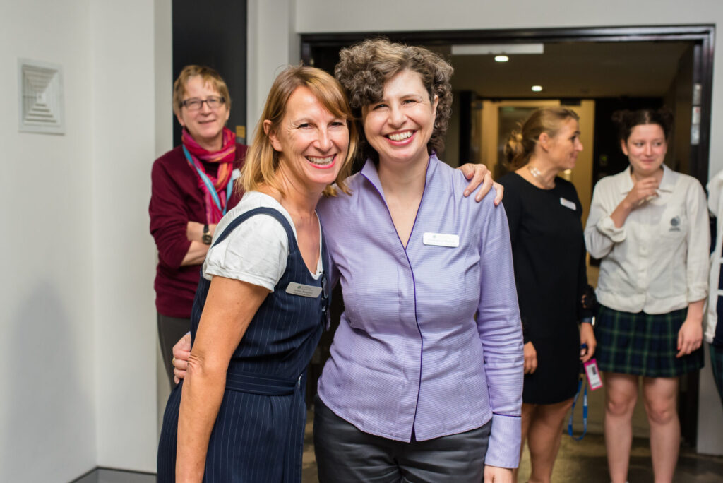 Two women stand smiling with arms round each other at an indoor IGS event, both wearing name badges. Three other people are in the background, two women talking and one person looking towards the camera—ready for a world-ready future of designing together.