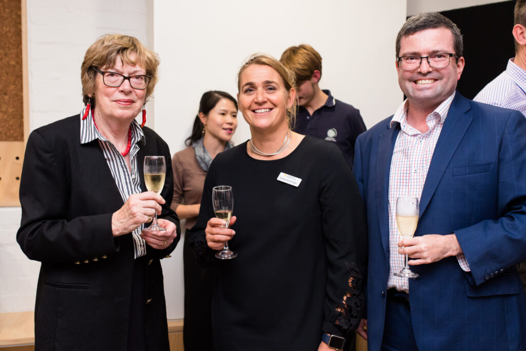Three people in formal attire hold champagne glasses and smile at an IGS event, surrounded by others engaged in conversation. The indoor setting suggests a world-ready reception or networking occasion, possibly focused on designing connections.