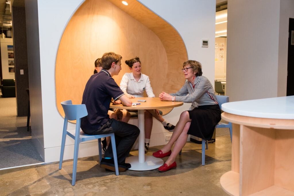 Four people sit around a round wooden table in a modern office space, engaged in conversation about designing world-ready solutions. Three appear to be IGS students, while one older woman in red shoes seems to be leading the discussion.