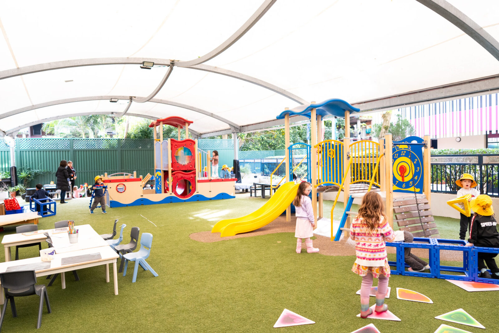 Children play on colourful playground equipment and engage in early learning activities at tables under a large white canopy in an outdoor play area, with adults supervising in the background.