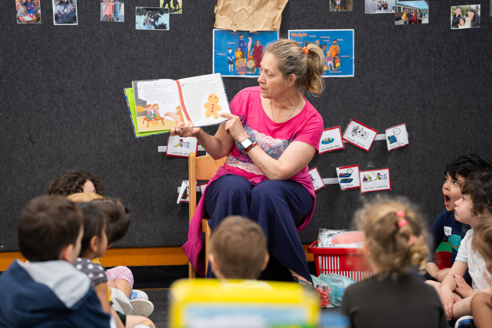 A woman sits in a classroom, engaging in Early Learning as she reads and shows a picture book to a group of young children seated on the floor. The children listen attentively, surrounded by colourful classroom decorations and photos on the wall behind them.