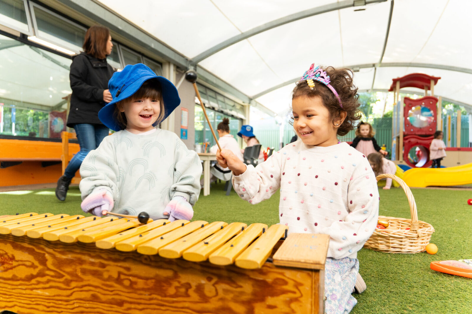 Two young children play a wooden xylophone together in a bright, indoor play area, fostering early learning. One wears a blue hat, the other a pink crown. Other kids and an adult are in the background near playground equipment.