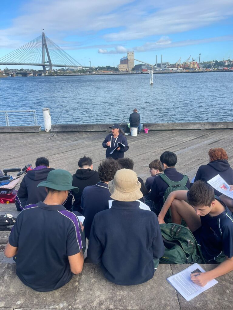 A group of Urban Explorers sit on concrete steps by the water, listening to a speaker. Behind them, a bridge, industrial buildings, and docks stretch across the river under a partly cloudy sky.