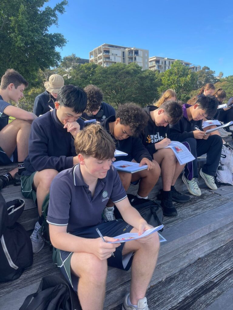 A group of Urban Explorers sits outdoors on wooden steps, focused on reading and writing in their notebooks on a sunny day. Trees and blocks of flats are visible in the background.