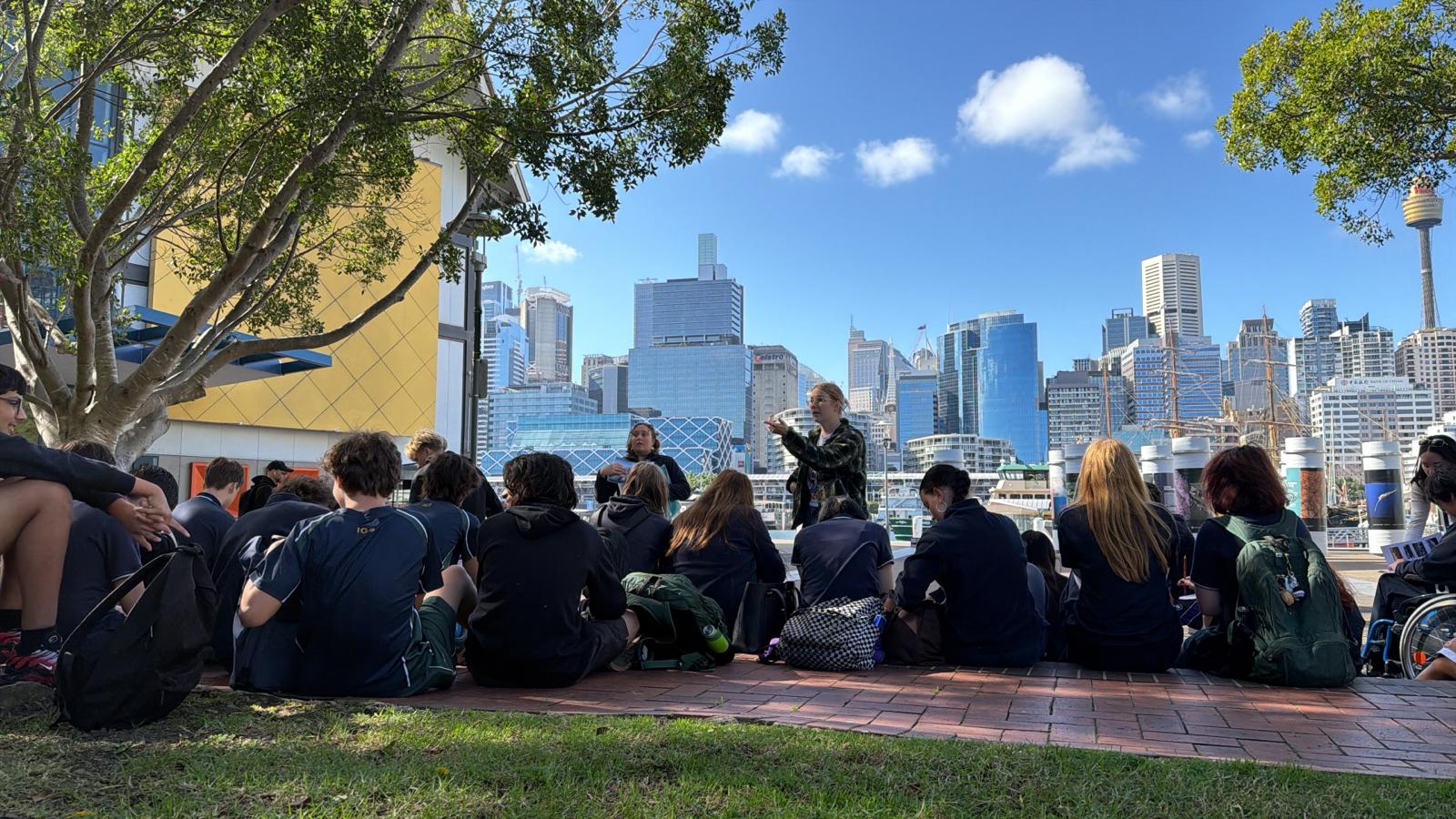 A group of Urban Explorers sits on the grass and brick path, facing a person speaking in front of them, with a city skyline and blue sky in the background. Trees and modern buildings complete the vibrant urban scene.