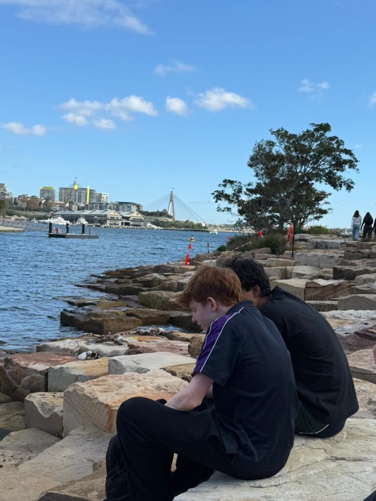 Two Urban Explorers sit on rocky steps by the water, facing away. In the background are trees, distant buildings, a bridge, and a blue sky with scattered clouds. Other people walk along the shore.