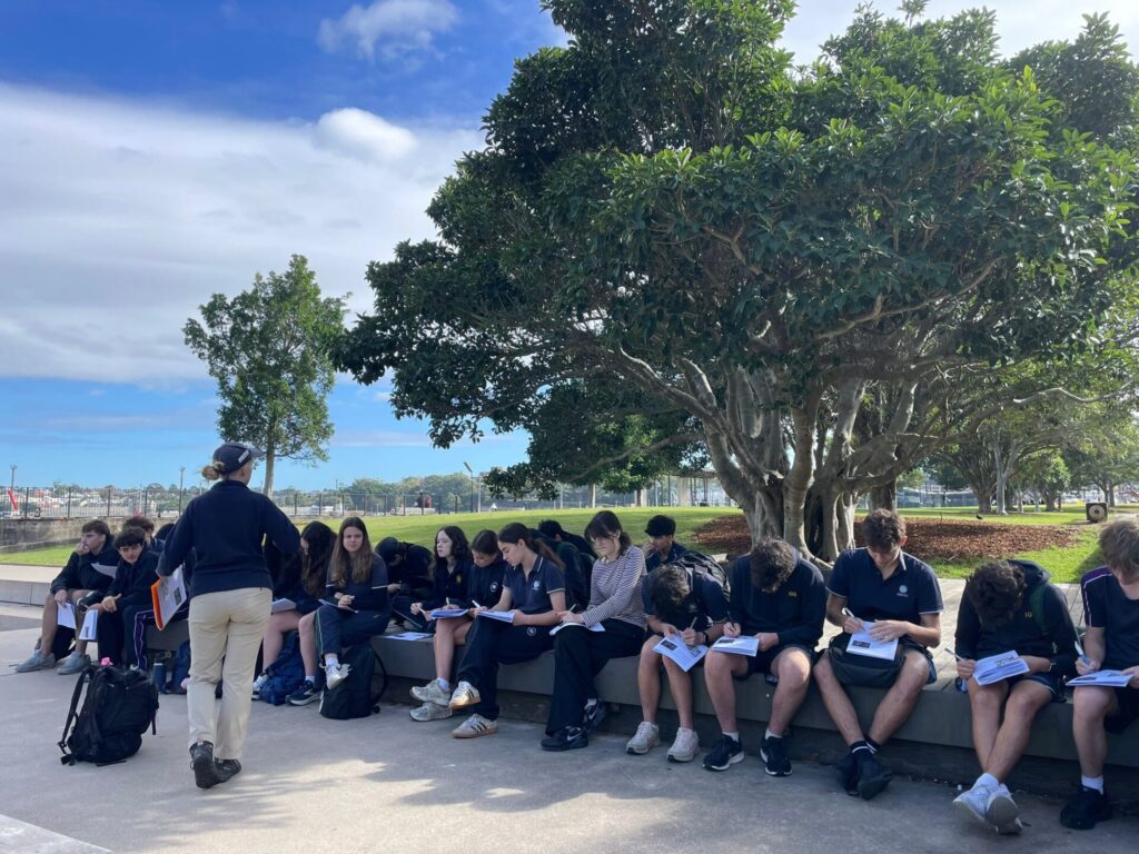 A group of Urban Explorers in uniforms sits on a low wall outdoors, writing in notebooks, while an adult stands nearby. Large trees and green grass surround them beneath a partly cloudy sky.
