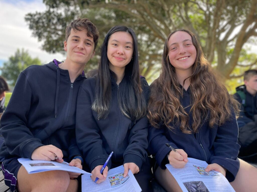 Three smiling teenagers sit outdoors on a bench, holding pens and worksheets, looking like Urban Explorers as they collaborate on a school project with trees and city life in the background.