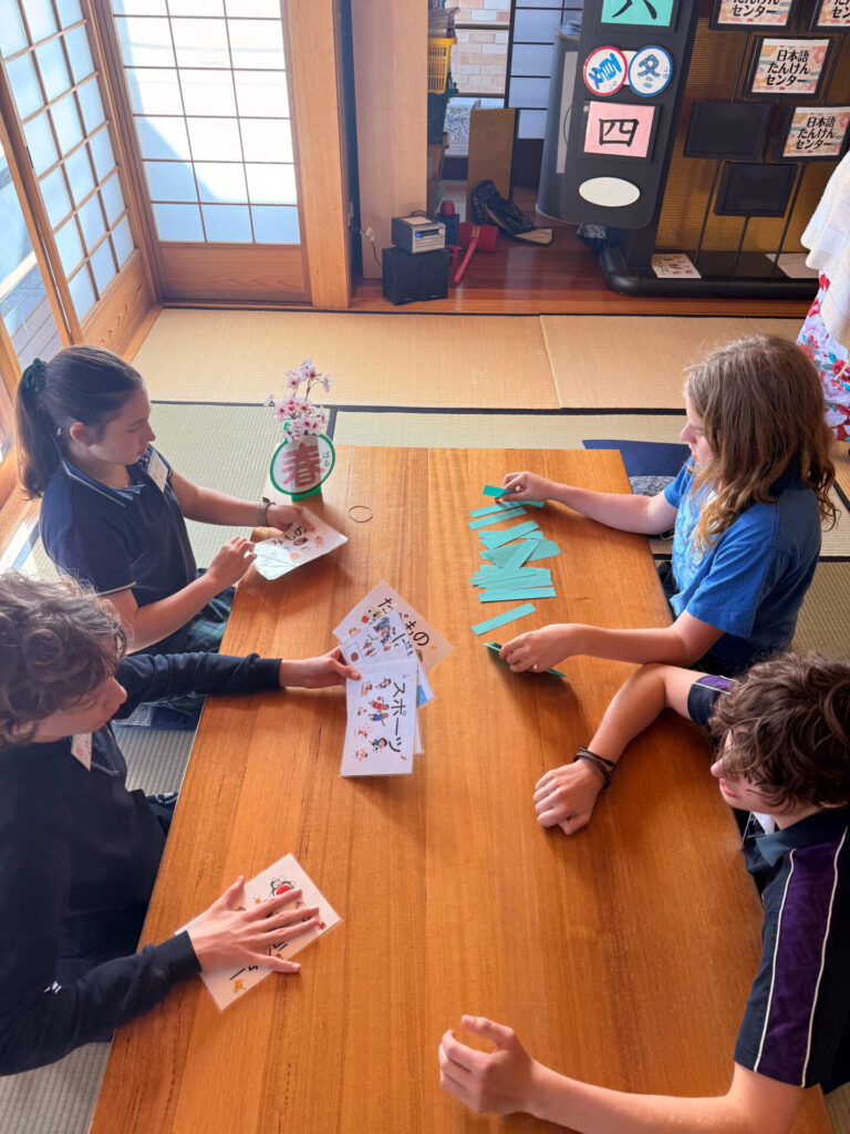 Four Year 9 pupils sit around a wooden table in a traditional Japanese-style room at the Nihongo Tanken Centre, playing a card game with green cards. Decorative items and Japanese calligraphy are visible in the background.