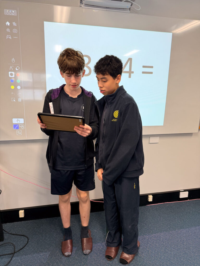 Two Year 9 pupils stand in a classroom; one holds a tablet while both look at it. Behind them, a projector displays a large maths equation, 3 + 4 =, on the whiteboard during their visit to the Nihongo Tanken Centre.