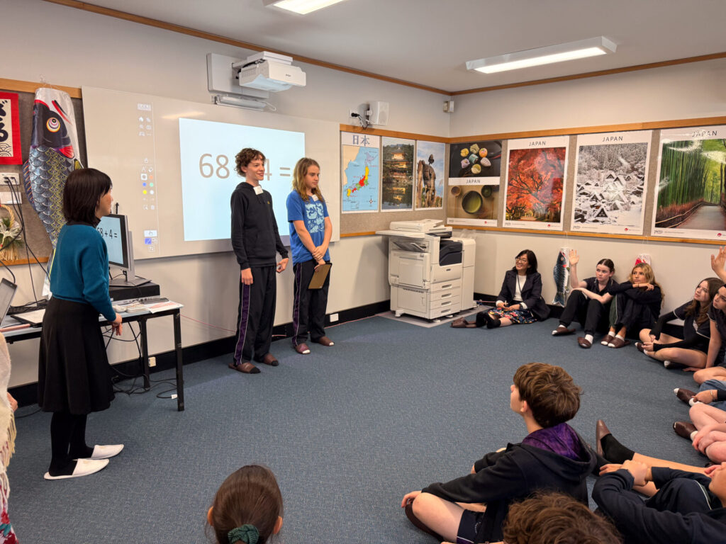 Year 9 pupils sit on the floor in a classroom at the Nihongo Tanken Centre while three classmates present in front of a screen displaying 68. Posters and maps about Japan decorate the walls, and one teacher sits among the pupils.