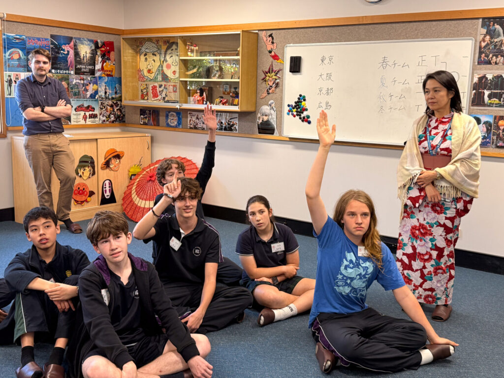 Year 9 pupils visit the Nihongo Tanken Centre, sitting on the floor—some raising their hands—in a classroom adorned with Japanese posters. A man stands by a cupboard while a woman in kimono teaches near a whiteboard with Japanese writing.