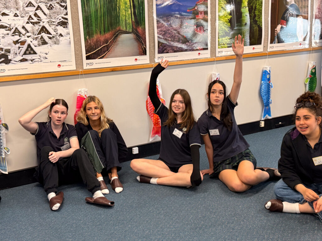 Five Year 9 pupils sit on a classroom floor in front of posters at the Nihongo Tanken Centre; two have their hands raised, others rest theirs on their heads or laps. They wear dark uniforms and socks, with colourful fish decorations hanging behind them.