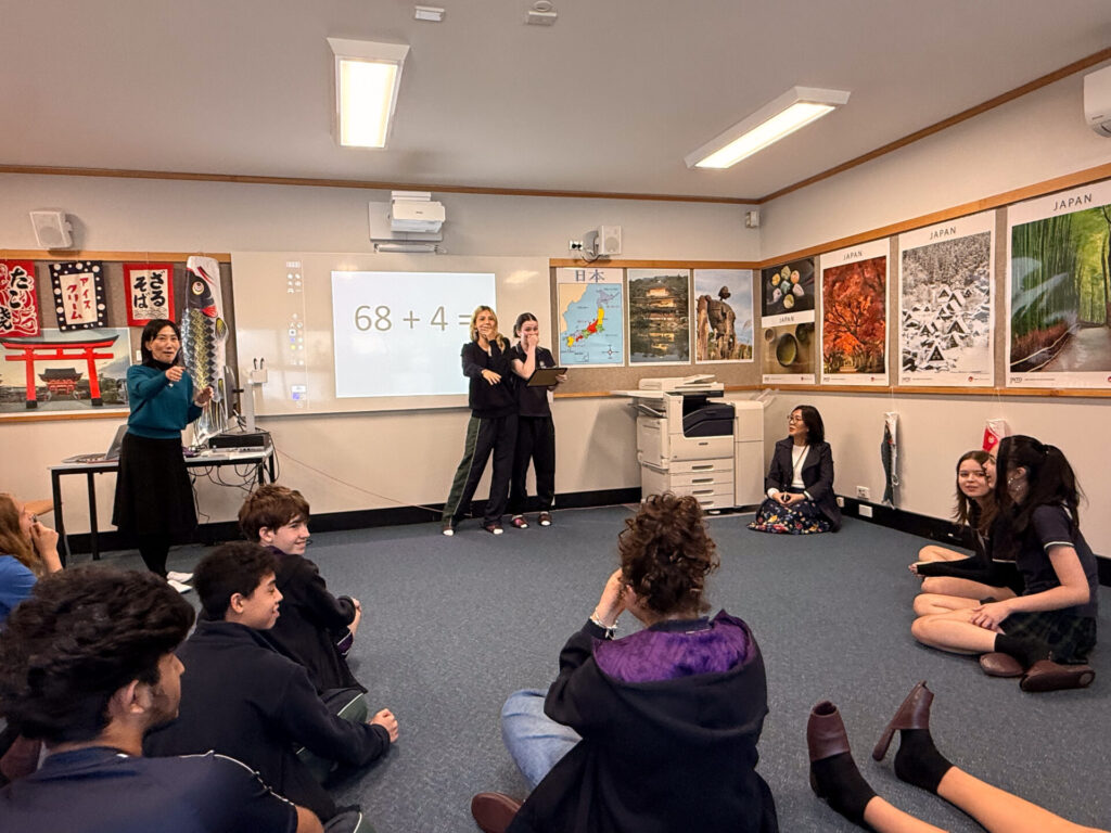 A classroom scene at the Nihongo Tanken Centre with Year 9 students sitting in a circle, two students at the front pondering “68 + 4 = ?” on a screen, surrounded by posters of Japanese culture and landmarks during their student visit.