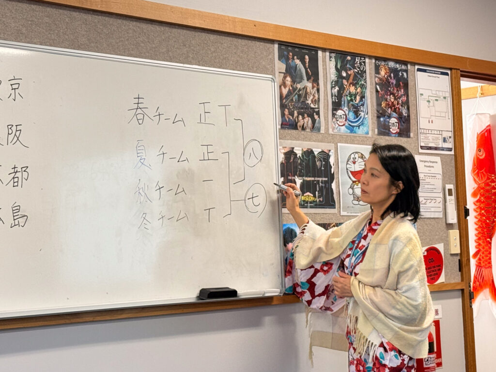 A woman in a kimono stands by a whiteboard with Japanese writing, explaining to Year 9 pupils at the Nihongo Tanken Centre. Posters and decorations are visible on the wall behind her.