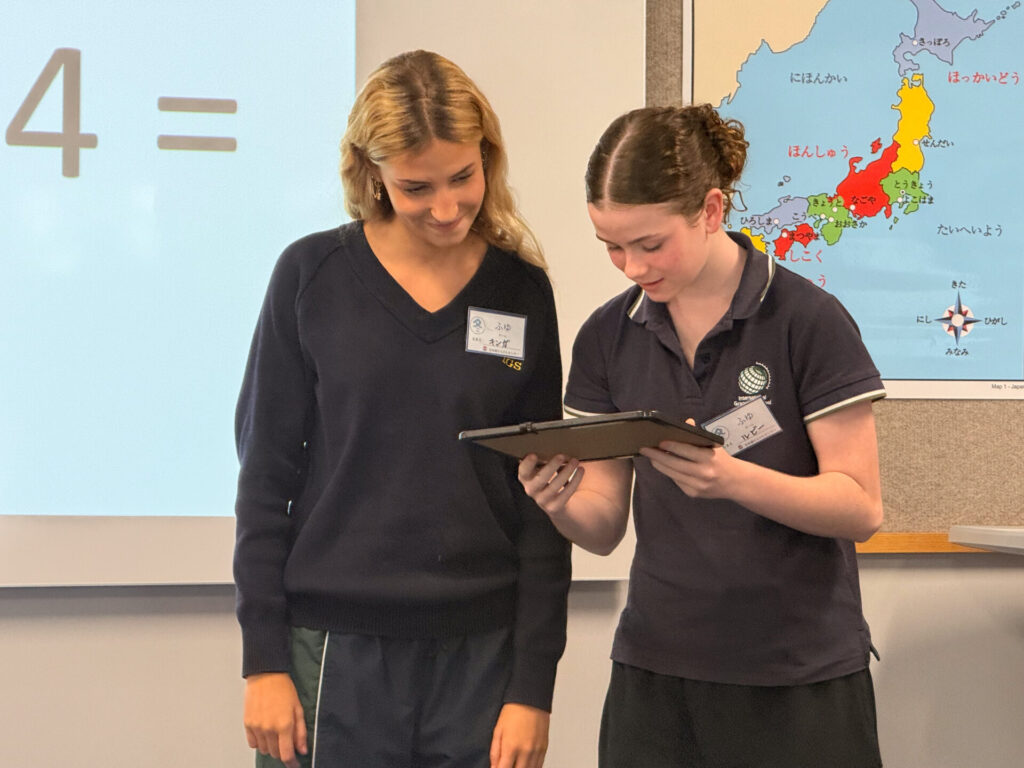 Two Year 9 pupils stand in a classroom at the Nihongo Tanken Centre, smiling and looking at a tablet together. A map of Japan and a whiteboard with large numbers are visible behind them as they explore the Japanese language.