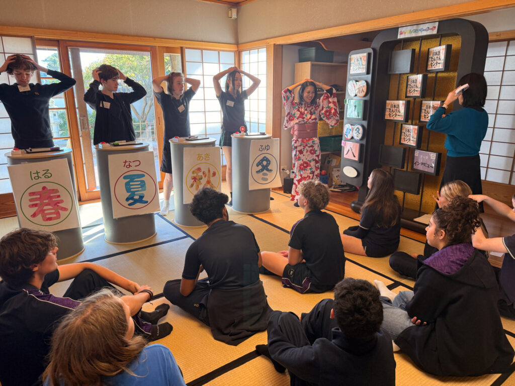 Year 9 pupils on a school trip to the Nihongo Tanken Centre sit on tatami mats watching a quiz game about seasons. Four participants stand at podiums as a woman in kimono leads, surrounded by Japanese decor.
