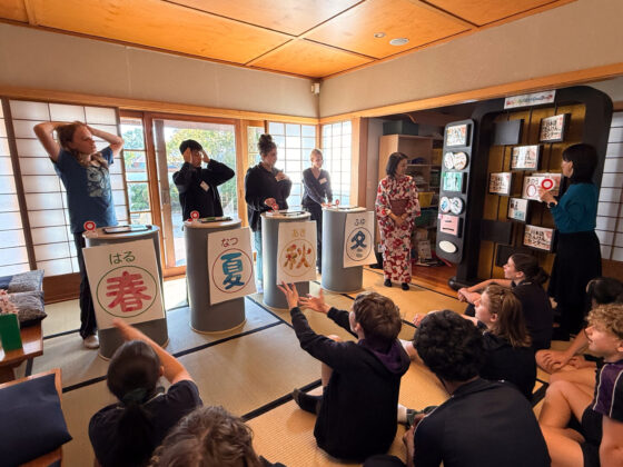 Year 9 pupils play a Japanese quiz game in a traditional tatami room at the Nihongo Tanken Centre. Four contestants stand behind season-labelled podiums, while others sit on the floor, raising their hands as a teacher moderates.