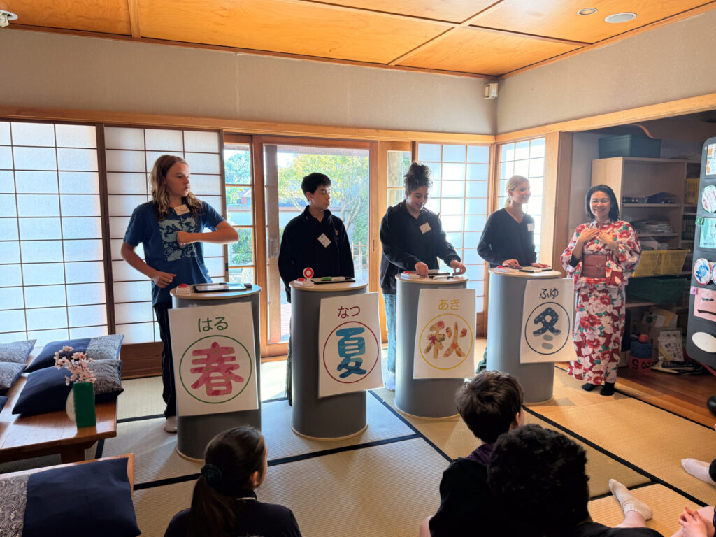 Five Year 9 pupils stand at podiums labelled with Japanese kanji for the four seasons, participating in a quiz game at the Nihongo Tanken Centre's traditional Japanese-style room, as an instructor in a kimono speaks to an audience.
