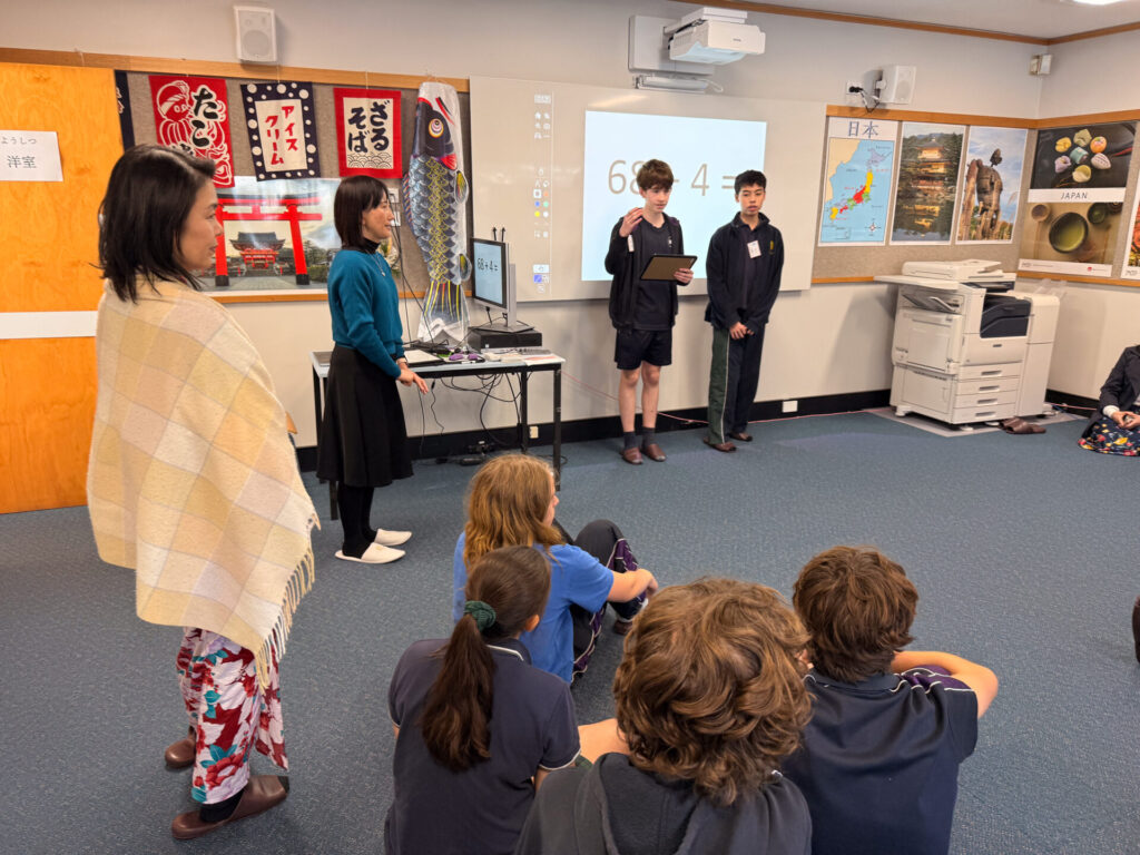 Two Year 9 pupils present in front of their classmates at the Nihongo Tanken Centre. Two teachers observe while Japanese posters and a map of Japan decorate the wall behind them. A timer is projected on the whiteboard.