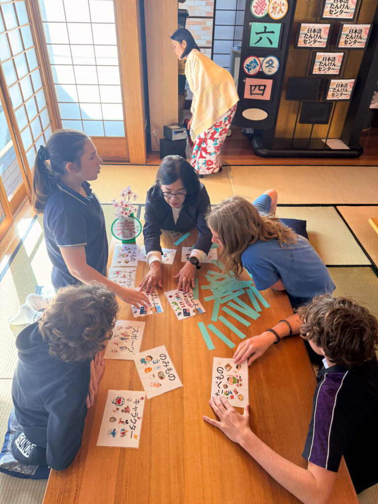 Year 9 pupils sit around a low table in a tatami room at the Nihongo Tanken Centre, engaging with Japanese language cards. An instructor in a kimono stands by a board with Japanese symbols, guiding their immersive school trip experience.