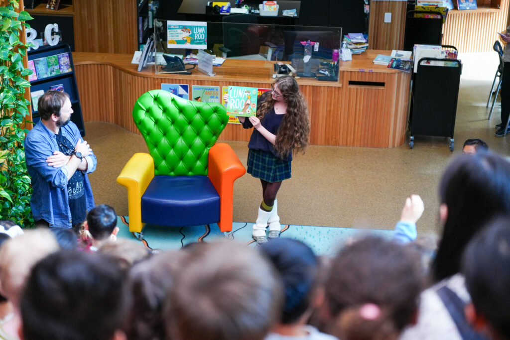 A woman stands beside a colourful armchair, holding up a book and reading to a group of children during storytime in a library setting. A man stands nearby, watching the early learning activity unfold.