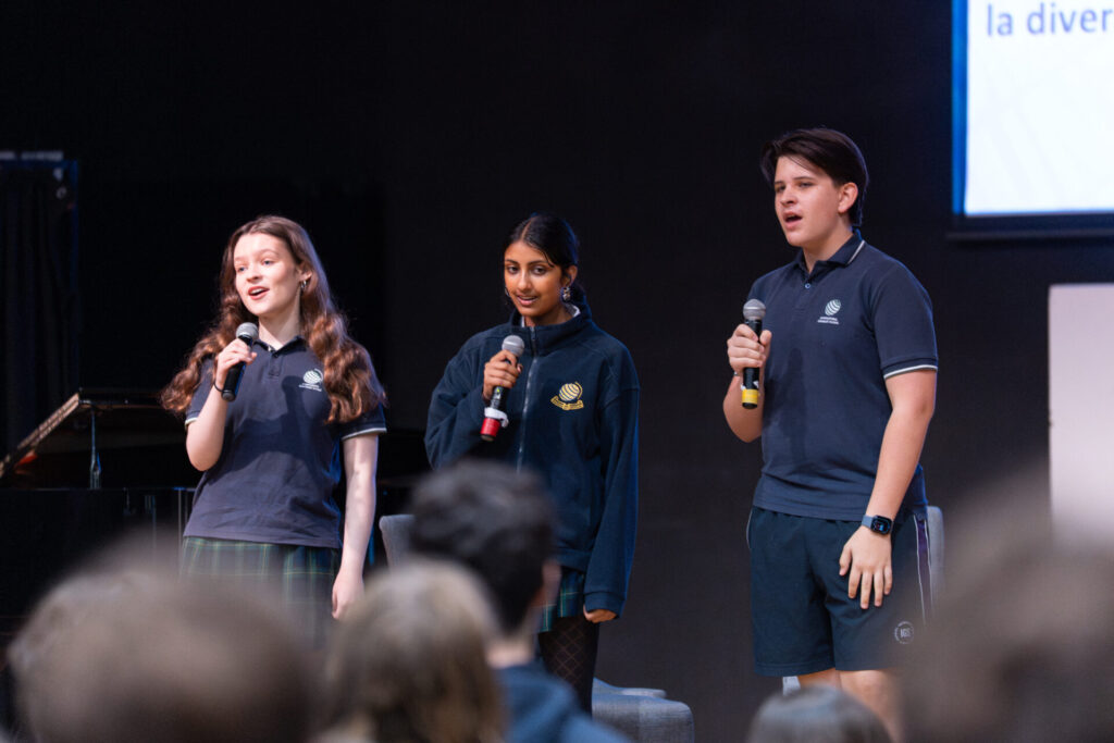 Three IGS students in school uniforms stand on stage holding microphones, singing or speaking to an audience during the Day of AI Australia event. A piano and screen hint at a bright future whilst the audience’s heads are slightly visible in the foreground.