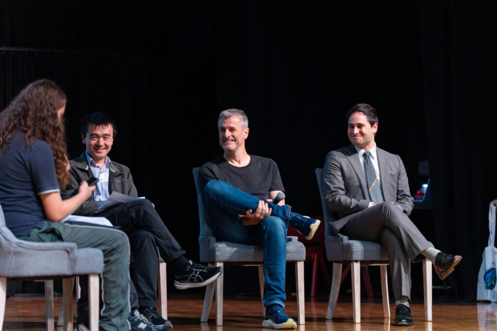 Four people are seated on stage in chairs, engaged in a panel discussion about artificial intelligence. Three men are dressed in varied attire, while a fourth person, seen from behind holding papers, speaks to them at Day of AI Australia.