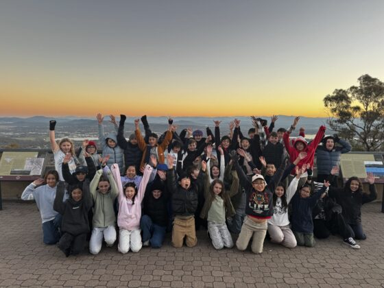 A group of excited children, dressed in warm clothing, pose outdoors at sunset with their arms raised, smiling widely. They are gathered on a paved lookout with panoramic views and informational signs behind them.